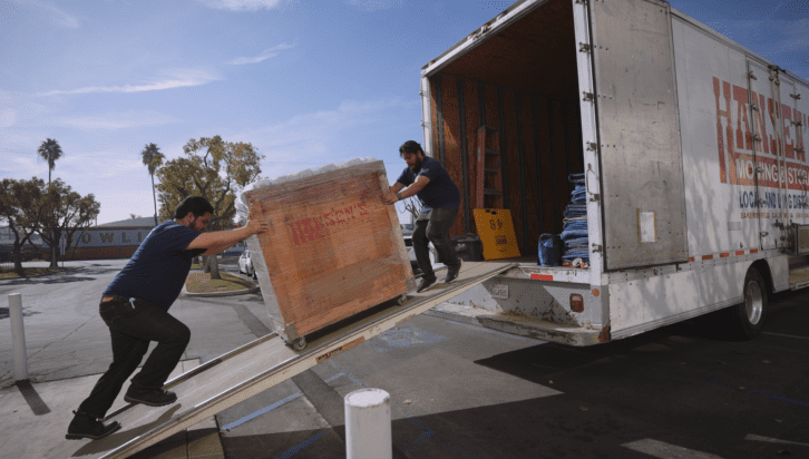 Professional movers are safely loading a large item into a moving truck using a ramp